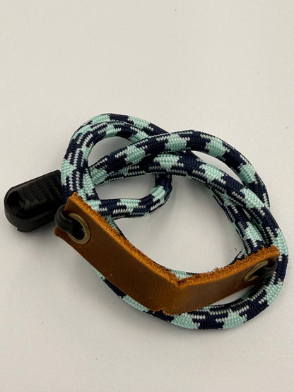 A coiled paracord EDC bracelet with a black plastic clip, featuring a navy, white, and light blue pattern, a brown leather basket, and brass grommets.